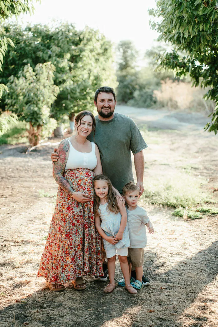A family of four standing together on a dirt path in a sunlit orchard