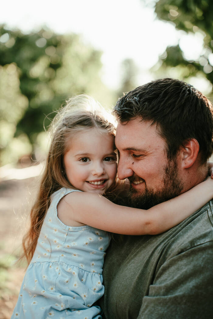 A young girl with her arms around her father, both smiling, backlit by warm evening light