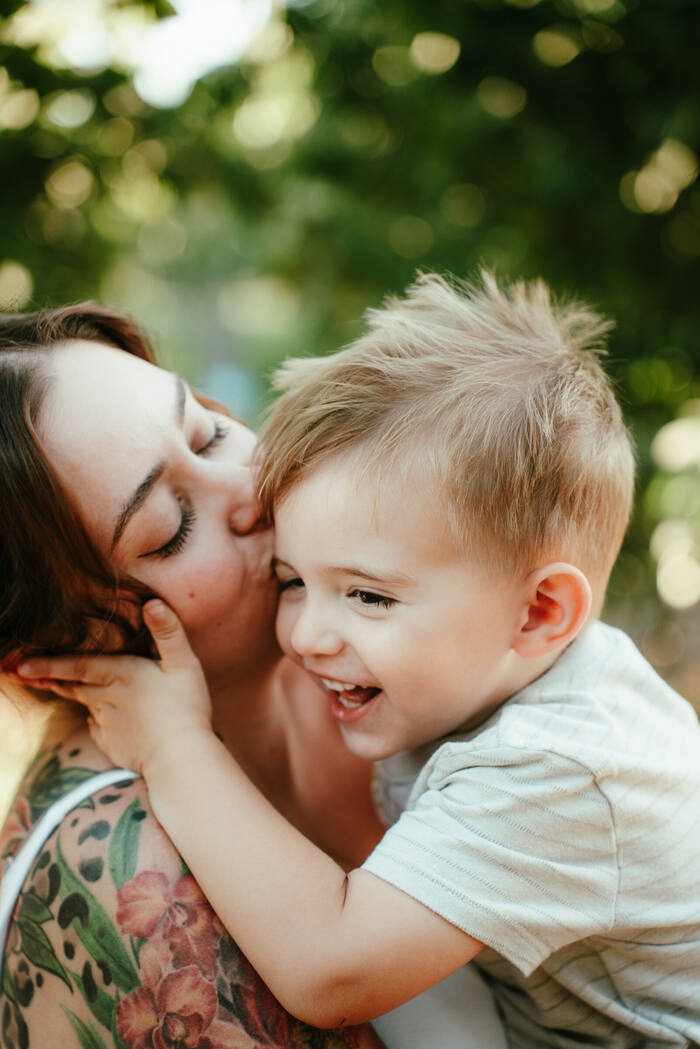 A mother kissing her laughing toddler son on the cheek, framed by green bokeh
